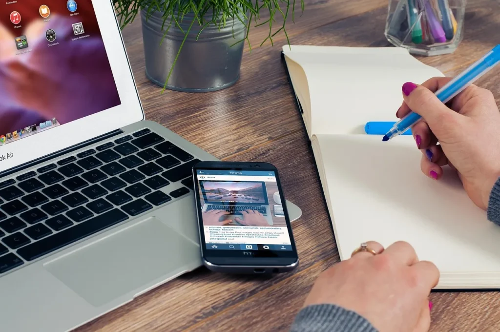 Bàn gỗ có laptop, điện thoại, sổ tay và bàn tay đang ghi chép, minh hoạ việc chuẩn bị hồ sơ xin việc sau tốt nghiệp